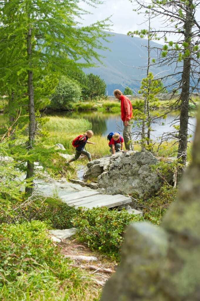 Familie wandert durch eine naturnahe Landschaft mit Kindern am Bachlauf und einem Erwachsenen.