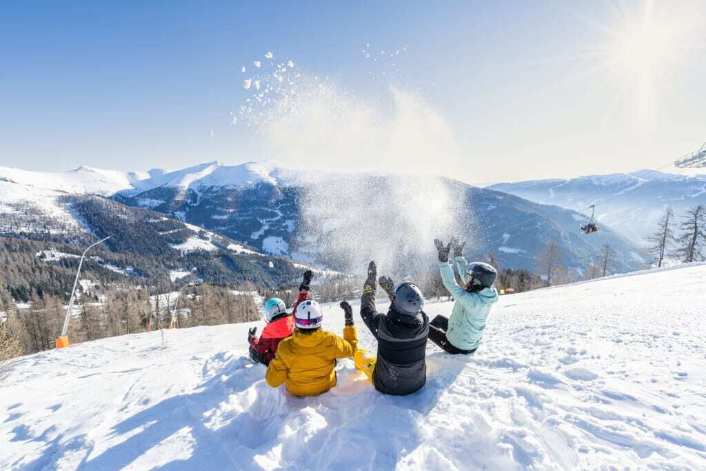 Gruppe von vier Personen auf einem verschneiten Hang, die fröhlich Schnee in die Luft werfen vor einer Bergkulisse.