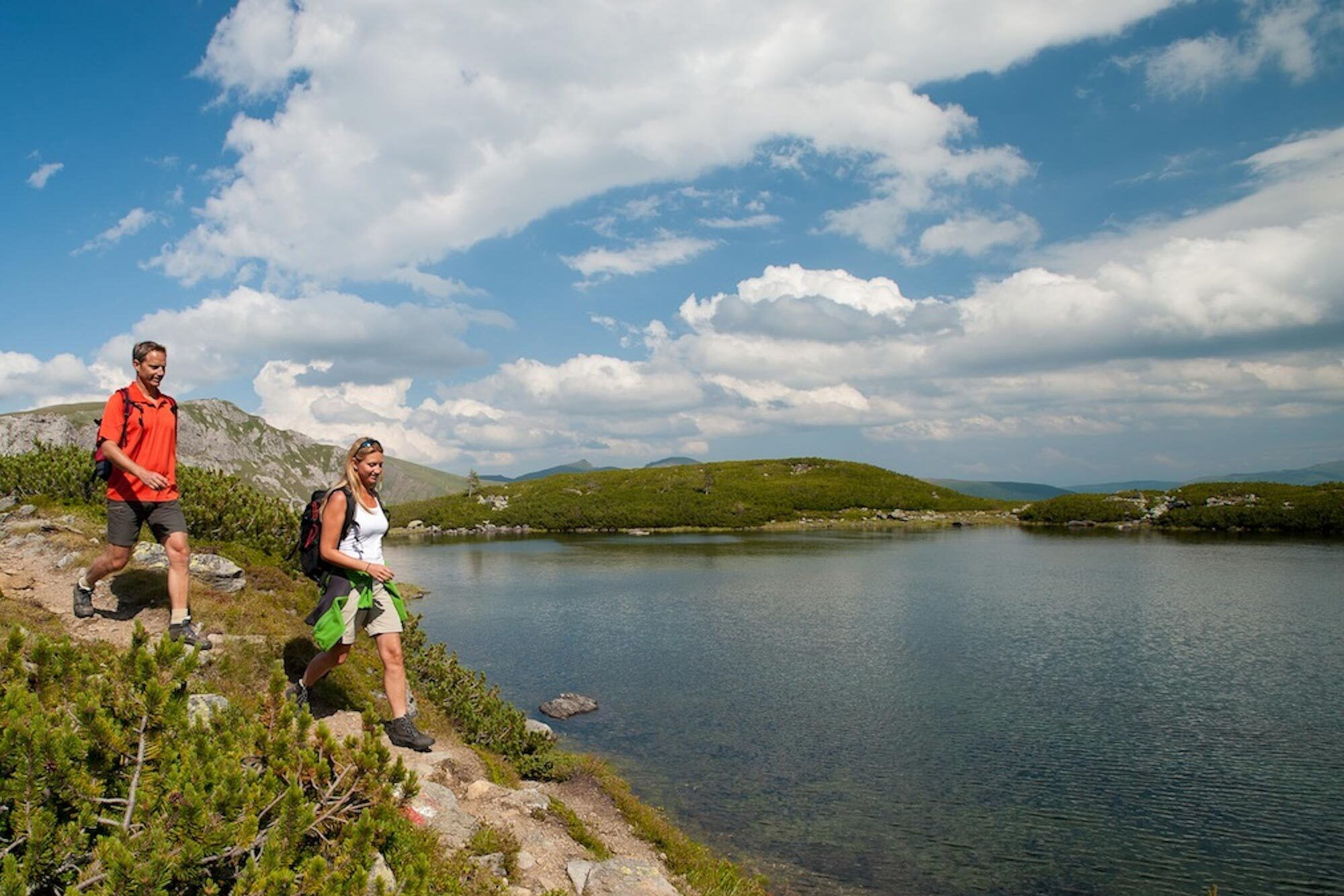 Zwei Wanderer auf einem Pfad am Ufer eines ruhigen Sees, umgeben von üppiger Natur und Bergen.