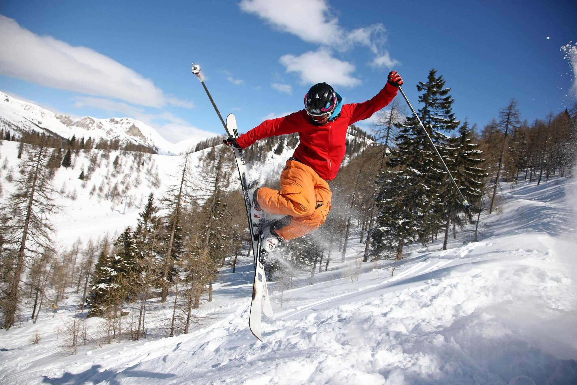 Ein Skifahrer springt über eine Schneepiste in einem Bergpanorama mit Bäumen im Hintergrund