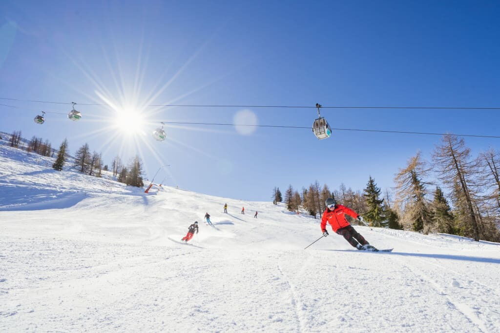 Mehrere Skifahrer auf einer schneebedeckten Piste unter blauem Himmel mit sichtbaren Skiliften im Hintergrund.