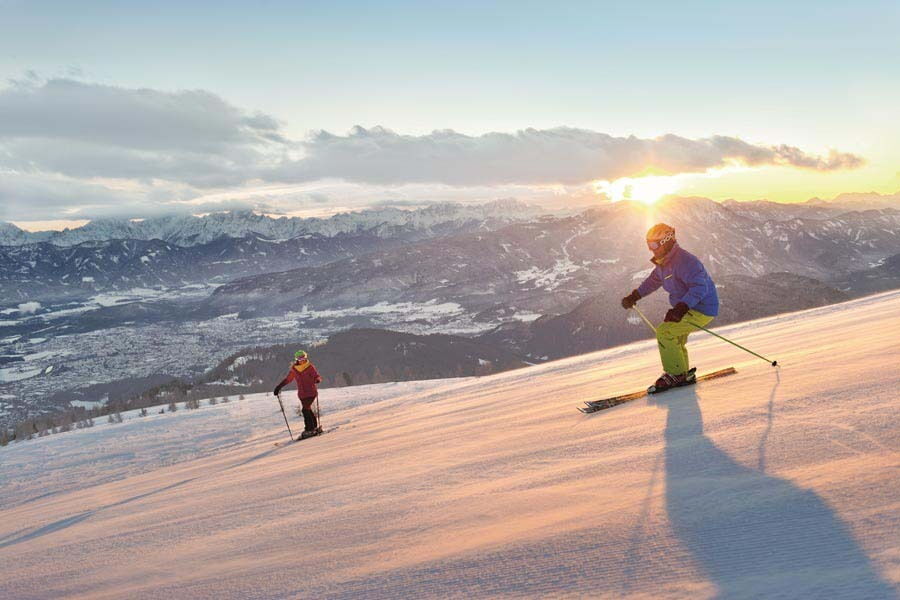 Zwei Skifahrer auf einer schneebedeckten Piste vor einem Bergpanorama bei Sonnenuntergang