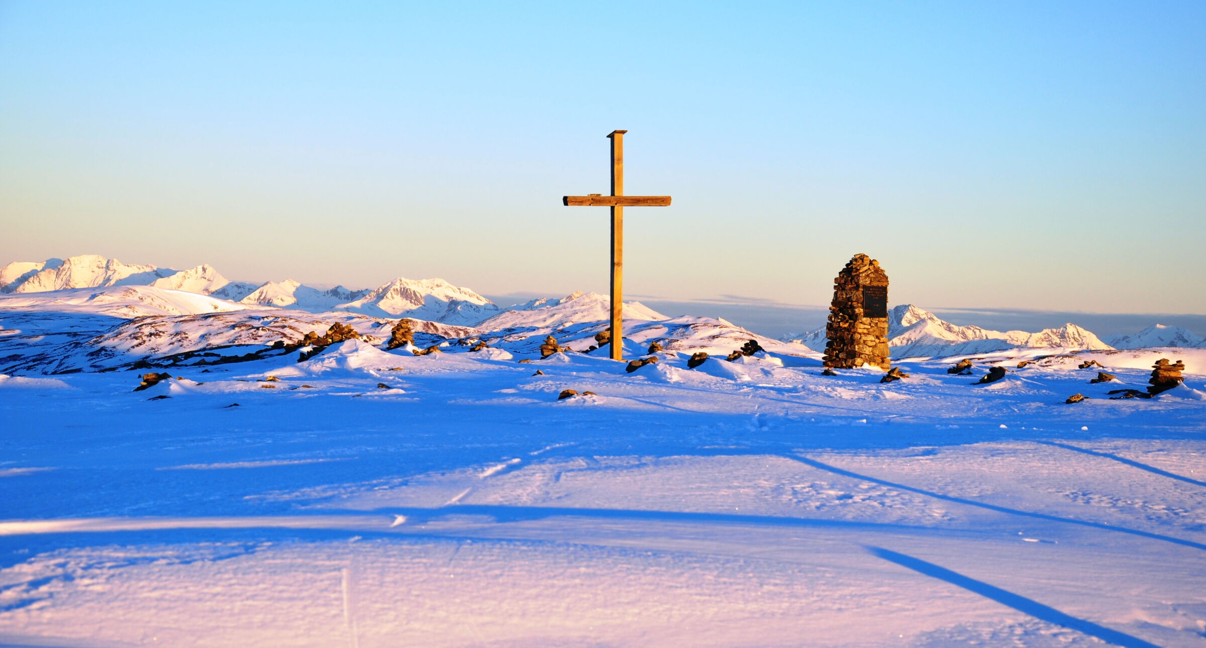 Schneebedeckte Landschaft mit Holz Kreuz und Steinsäule vor majestätischen Bergen.