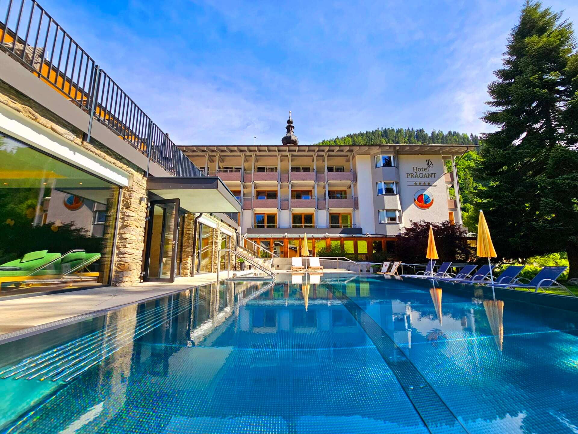 Hotel mit Swimmingpool und Berglandschaft im Hintergrund, umgeben von grüner Vegetation.