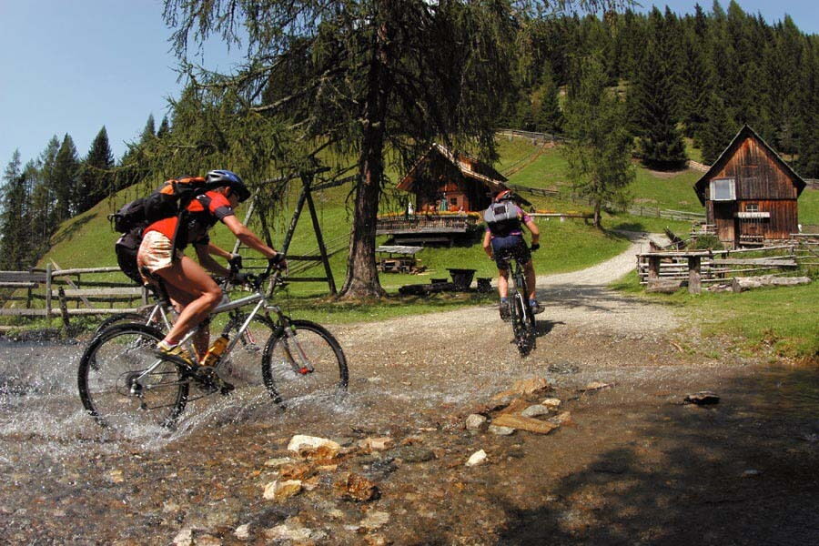 Zwei Mountainbiker fahren durch einen Bach in einem Waldgebiet mit Hütten im Hintergrund