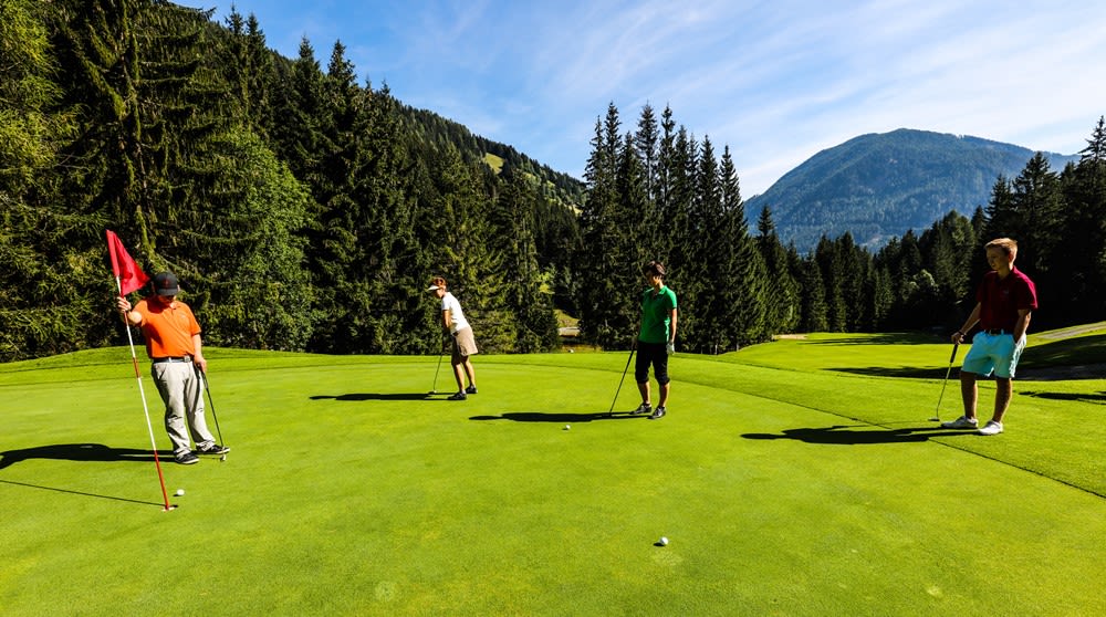 Vier Personen spielen Golf auf einem gepflegten Platz, umgeben von Bäumen und Bergen.
