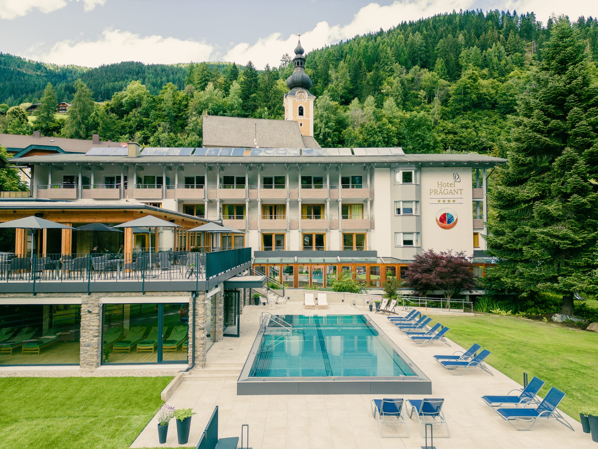 Hotel mit Schwimmbad und Liegen vor Berglandschaft und Bäumen im Freien gelegen.